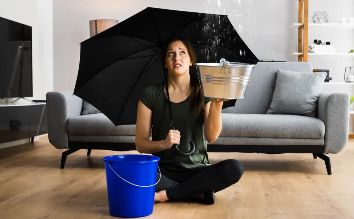Mulher sentada na sala segurando guarda-chuva e balde sob uma goteira no teto